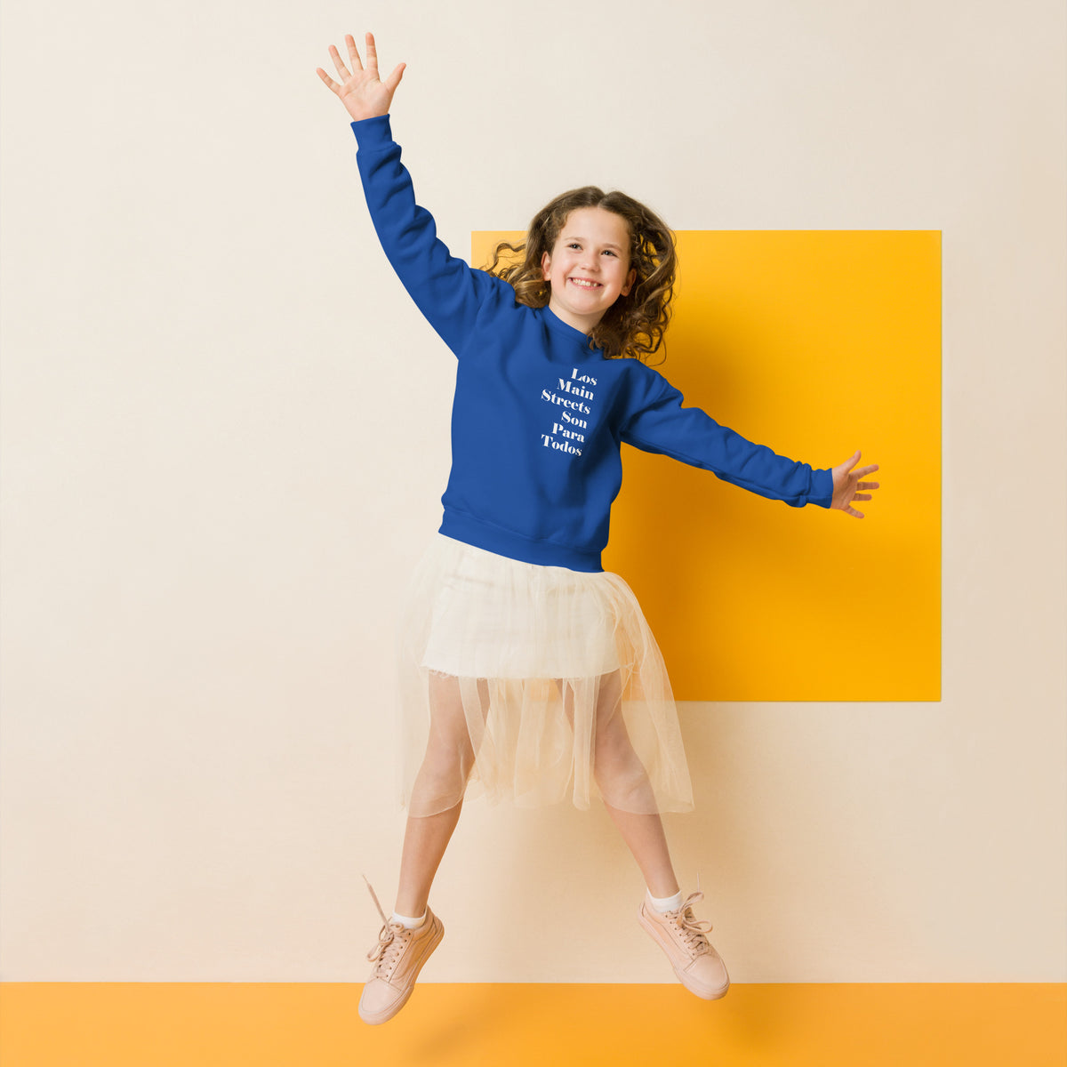 Child wearing blue sweatshirt reading "Los Main Streets Son Para Todos" jumps with arms outstretched in front of yellow and white wall